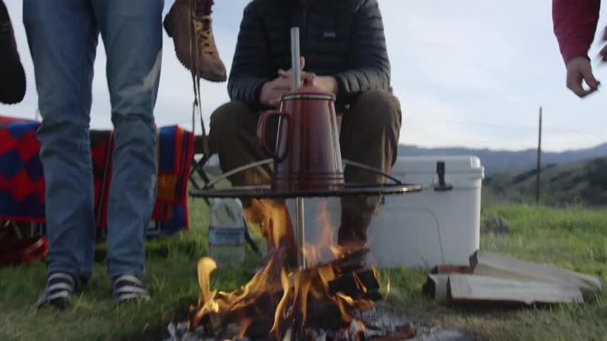 A man holds a boot over a campfire as coffee is being made