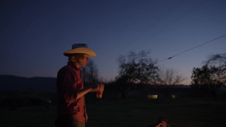 A group of friends sits around a campfire and drinks beverages in the field at dusk