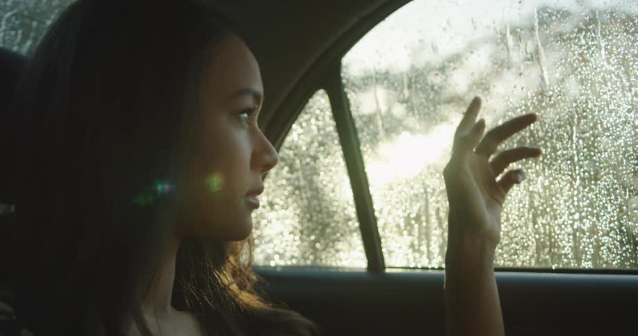 A young woman sitting in the backseat of a vehicle on a rainy day fidgets with her hand as she sits lost in thought