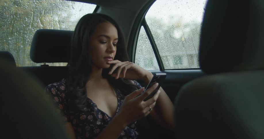 Woman with straight dark hair and a dress sits in the back seat of the car and uses her smartphone as it rains outside