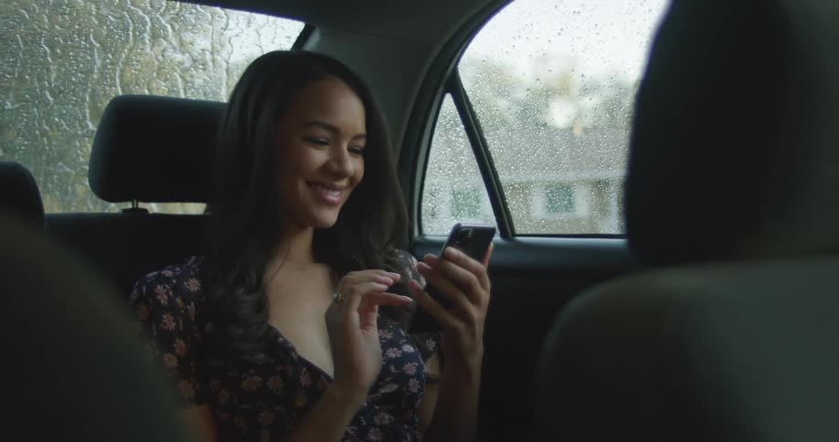 A teenage girl sits in the back seat of a car and laughs as she looks at her cell phone