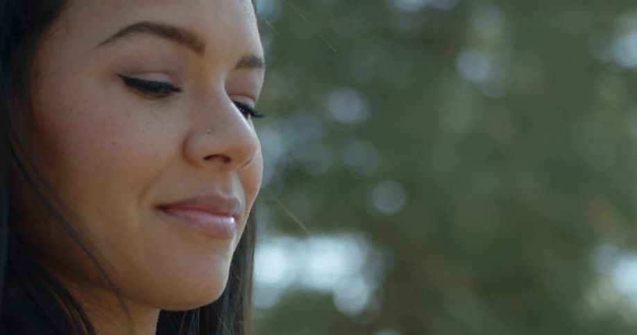 Brunette woman with dark straight hair sits on a swing under the tree and smiles slightly