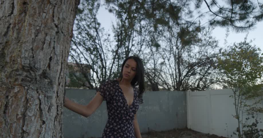 Brunette woman with a dress touches and walks around a tree in the backyard