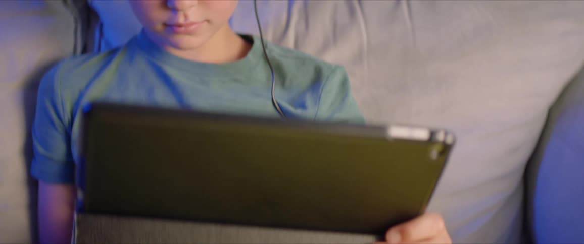 Boy with a green shirt and headphones sits on sofa and uses tablet in the living room at home