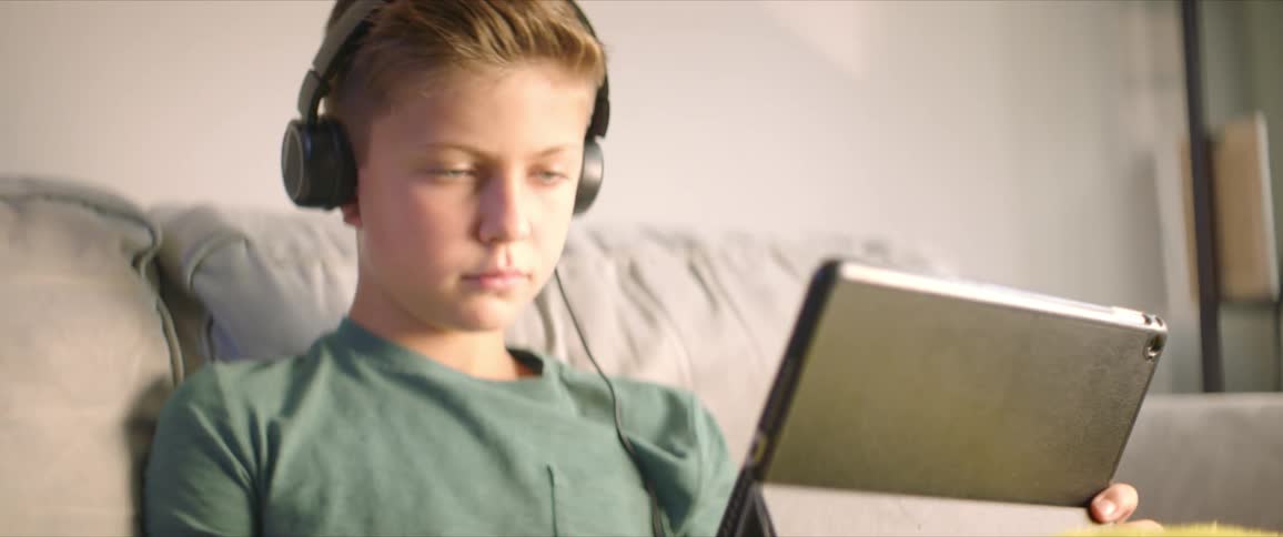 Boy with black headphones and a green shirt sits on the sofa and writes in a notebook while using a tablet in the living room at home