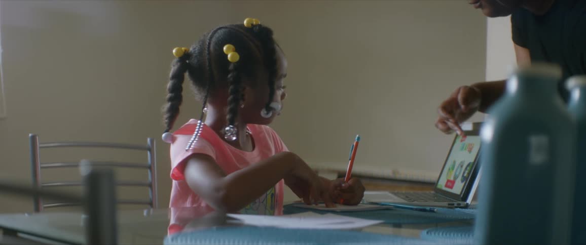 A young girl sits at a table writing and a woman approaches to help with something on the tablet screen in front of her