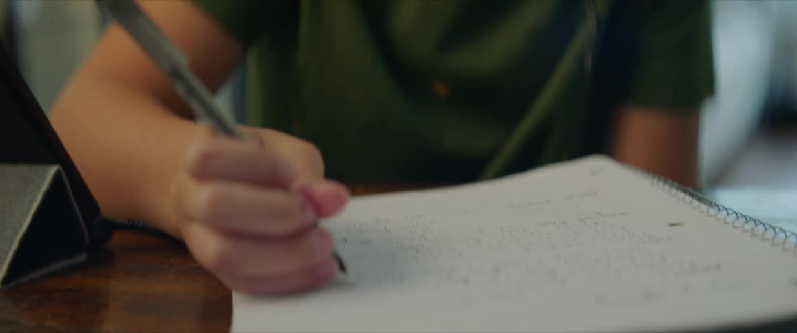 A young boy sits at a table wearing headphones and writing in a notebook with a tablet next to him