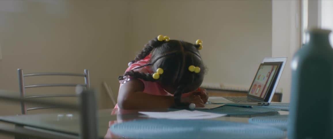 A woman walks over to check on a young girl sitting at a table with a tablet and writing on a piece of paper