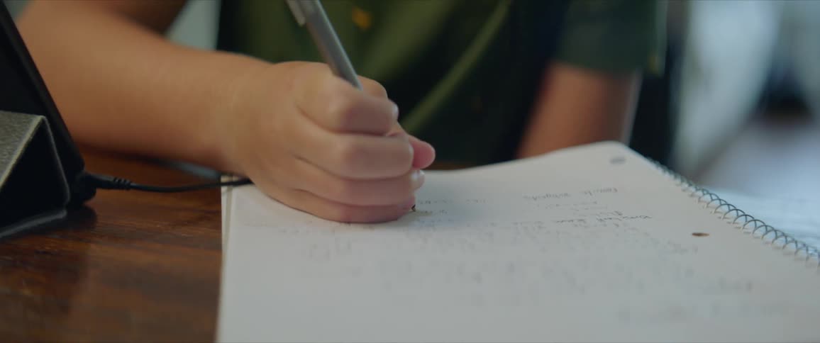 A young boy sits at a table writing in a notebook and listening to a tablet with headphones