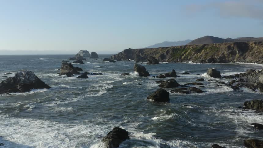 Rock formations and boulder poke through the tide off the coast of a landmass during the day