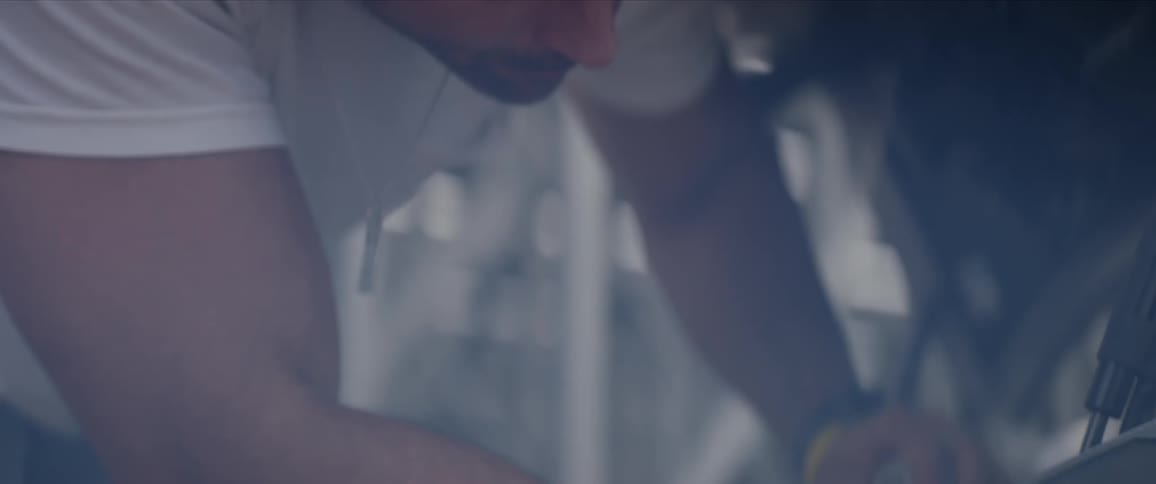 A man wearing a white t shirt reaches into a car and wipes of a tool with a towel as he nods his head