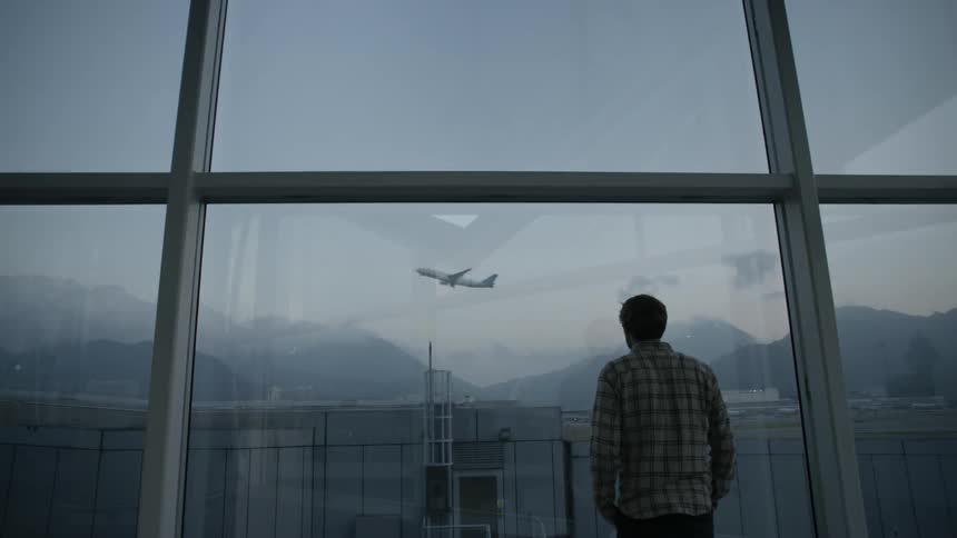 A man watches through large windows at an airport as an airliner takes off