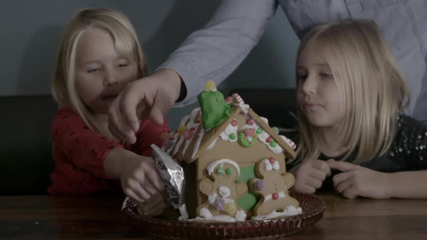 Two children are working on a gingerbread house as an adult helps