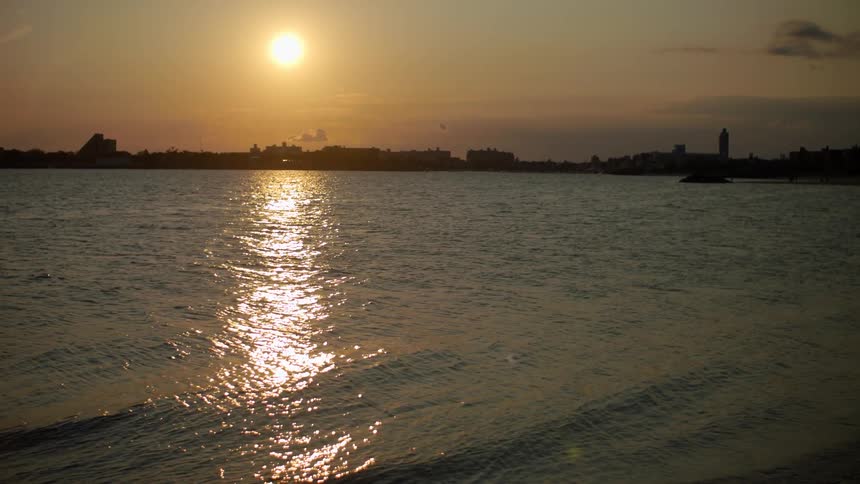 Sun sits in red sky glaring off ocean washing onto shore near silhouetted skyline