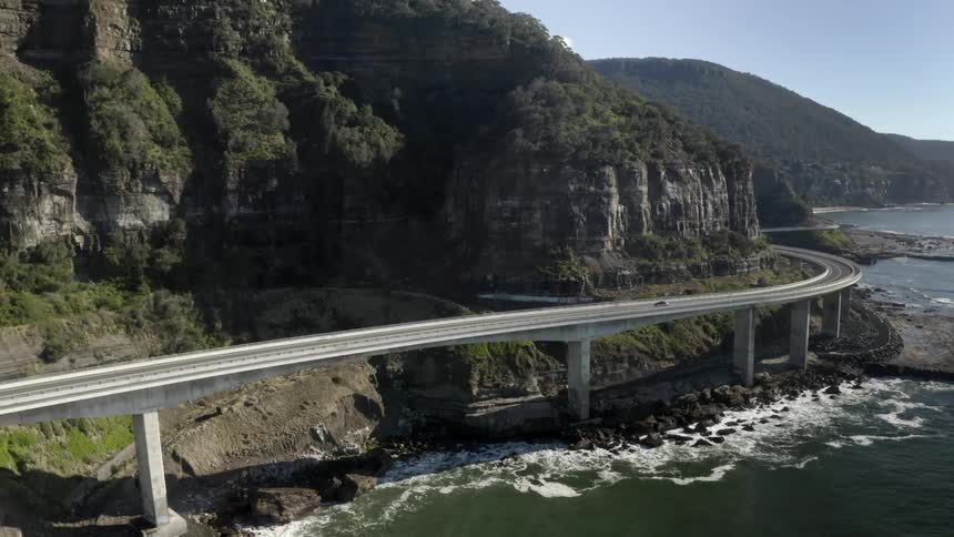 Cars drive on an elevated highway above the ocean at the coastline next to a hill in daylight