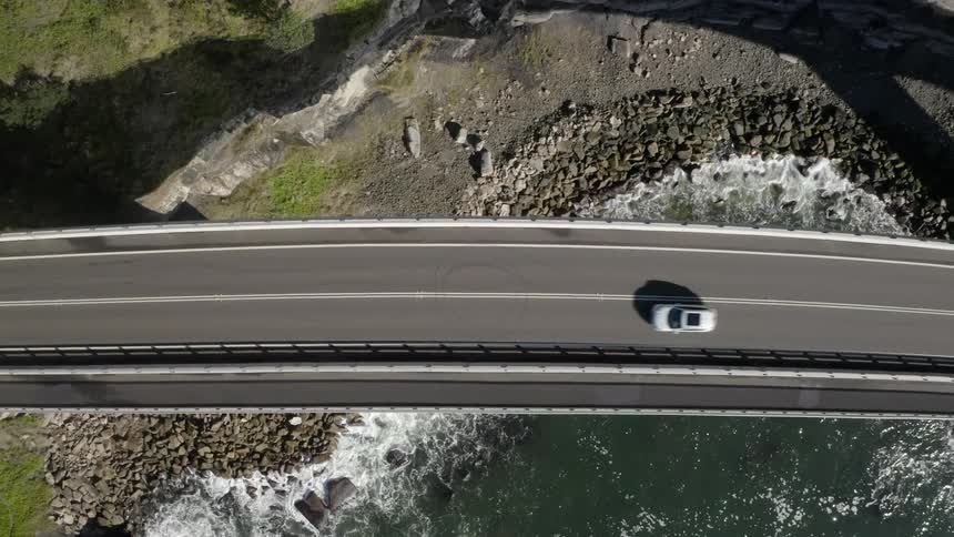 A vehicle travels across a bridge over a cove with a rocky beach during the day
