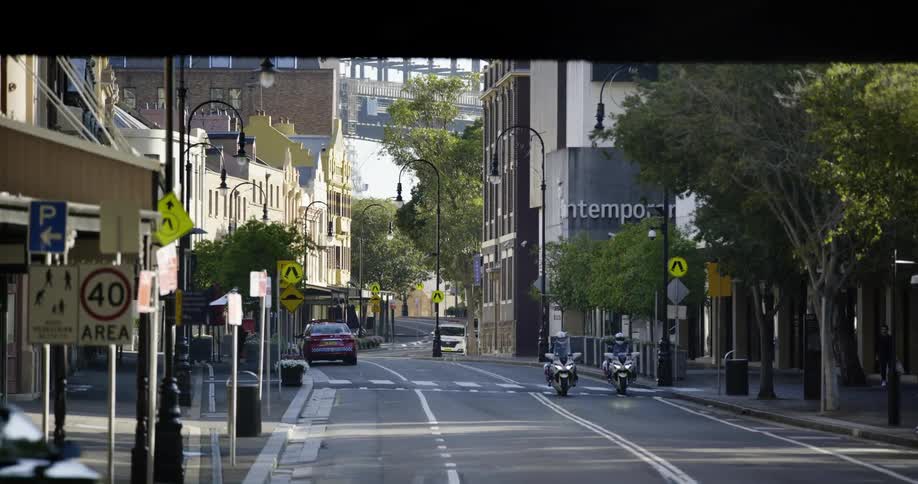 Two people drive motorcycles down a city street with a car and another motorcycle driving on the other side of the road on a sunny day