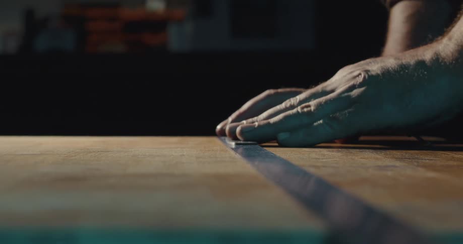 Pair of hands taking measurements and placing a strip on top of a wooden board inside of a workshop