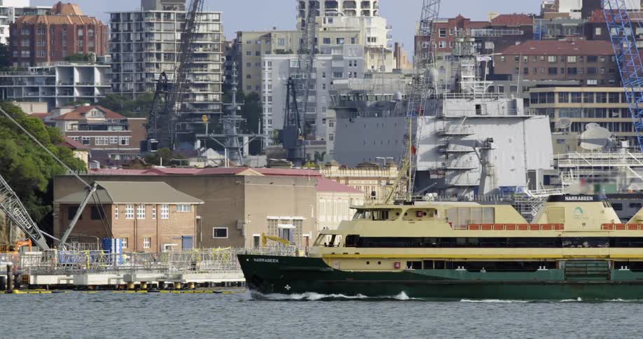 A green and yellow ferry moves along the water in front of a coastline with a city