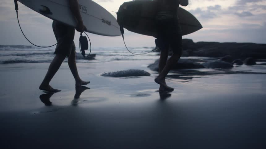 Two men carry surfboards while walking down a beach with rocks