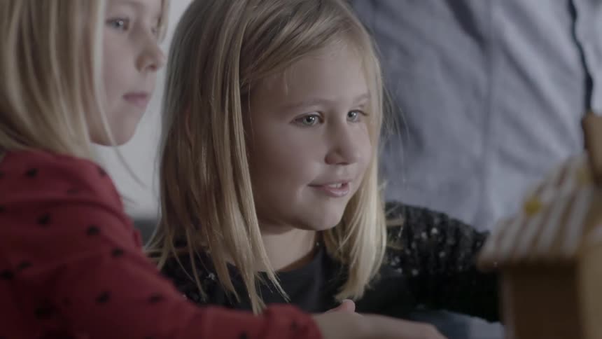 Two girls watch someone sprinkle something on a gingerbread house