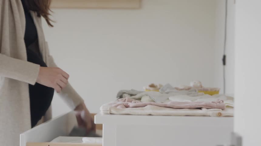 Young long haired pregnant woman folding baby clothes over a dresser in a room at home