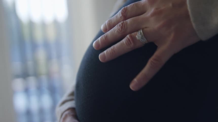 Pregnant woman with a black sweatshirt and rings touches her belly in a room