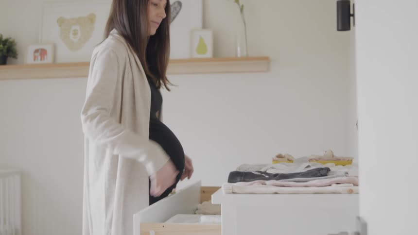 Pregnant woman walks into a baby room during the day and picks up baby clothes on top of a dresser to fold them