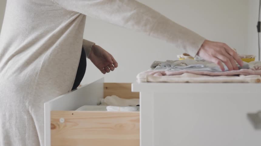 Pregnant woman standing in a nursery folding baby clothes and placing them into a dresser