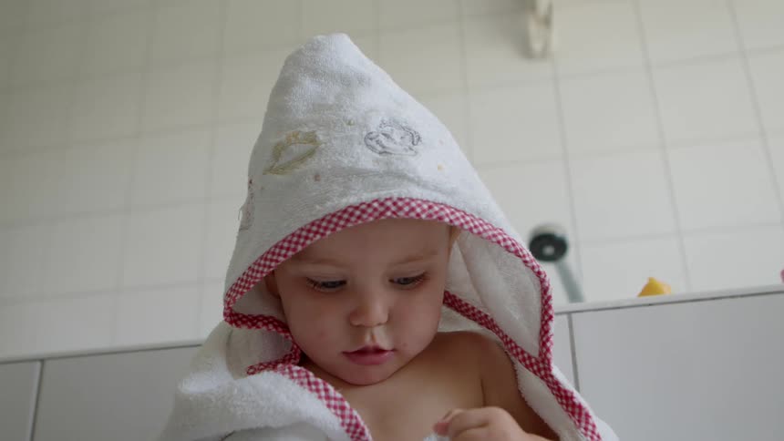 Baby wrapped in a bath robe sitting on the floor of the bathroom next to the bathtub