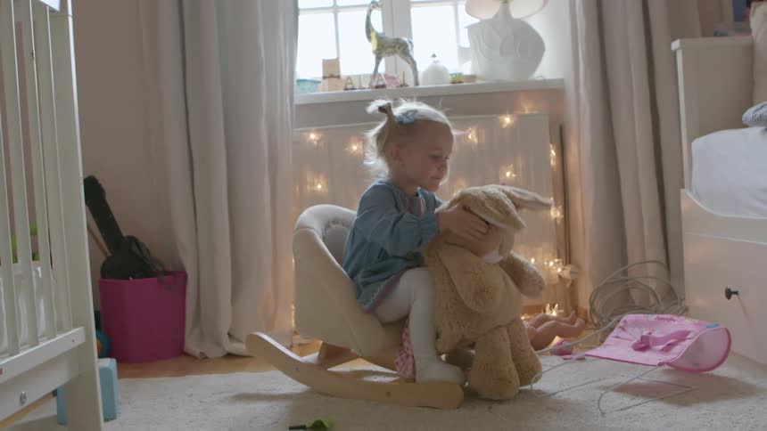 Little girl sitting in a rocking chair playing with a stuffed bunny in her room
