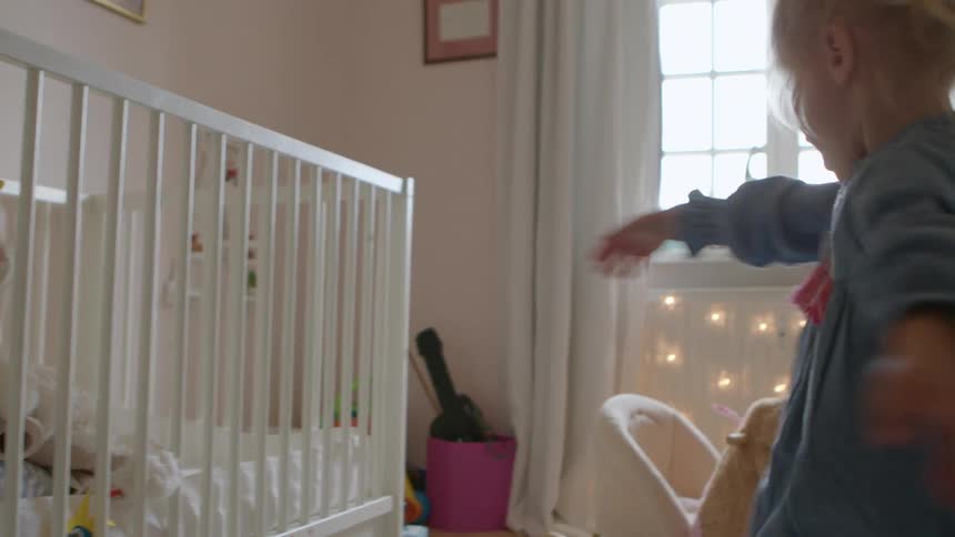 A young girl standing near a crib in the bedroom of a house spins around in a circle before falling over