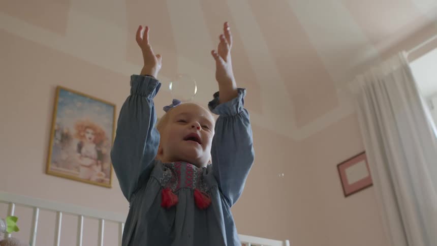 Little girl in a pink and white striped childs room playing with bubbles being blown
