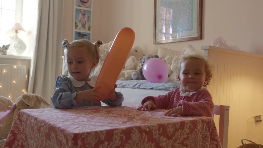A pair of young girls play with balloons while sitting at a small table in a childs bedroom during the day