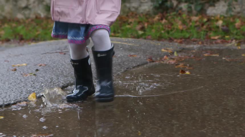 A young toddler girl runs in circles in a puddle wearing rain boots