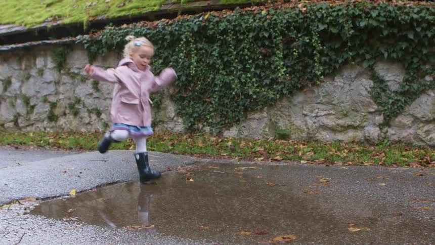 Little girl with a pink coat and black boots jumps over a puddle