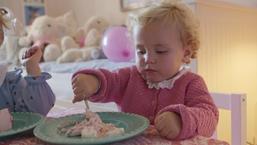 A blonde toddler plays with a piece of cake while sitting next to another girl at a table indoors during the day