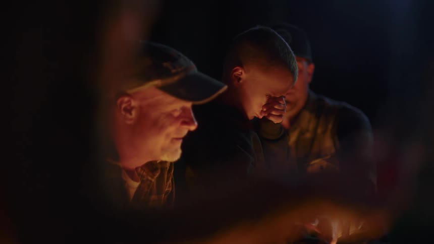 Boy standing up outside at night with his father and grandfather poking a fire with a stick