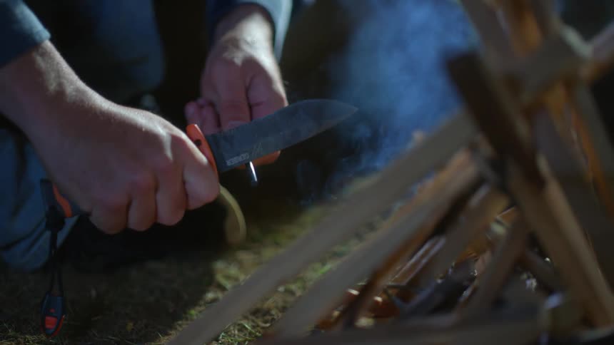 A hand holds flint while the other hand strikes a flint creating a flame in front of wood