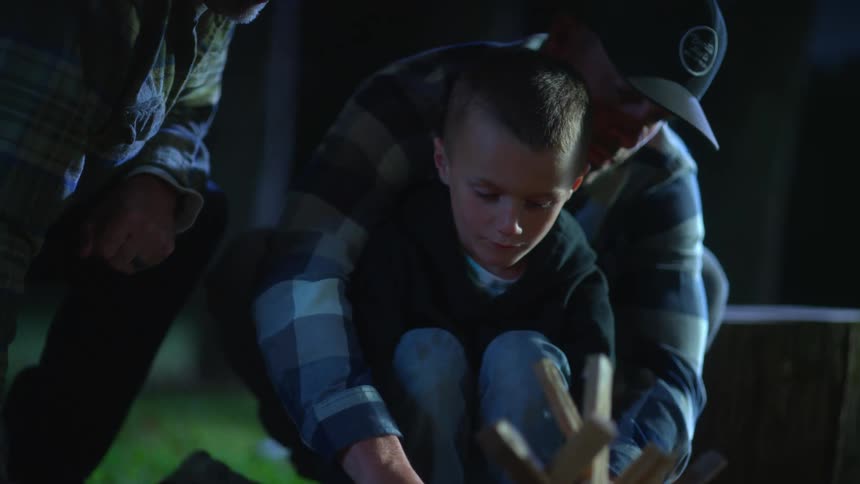 Father teaching his son how to light a fire outdoors while another man watches on at nighttime