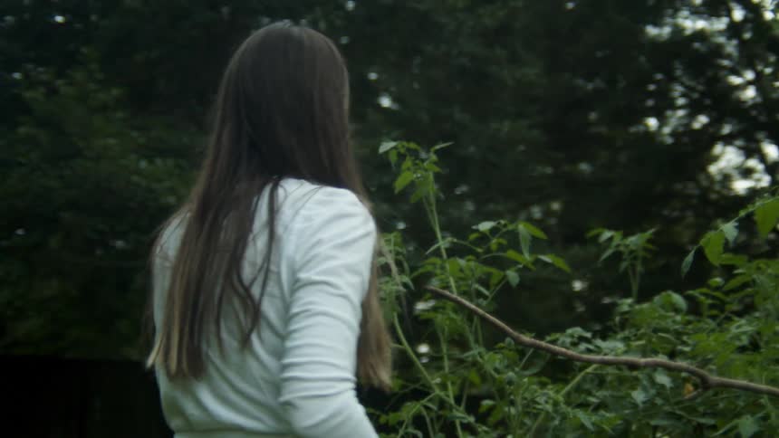 A woman standing next to a plant picking berries with trees the sky and clouds in the background