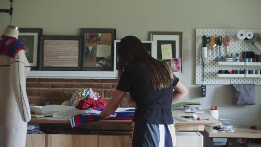 Woman inside of a sewing workshop standing in front of a table using scissors to cut through a blue and red shirt