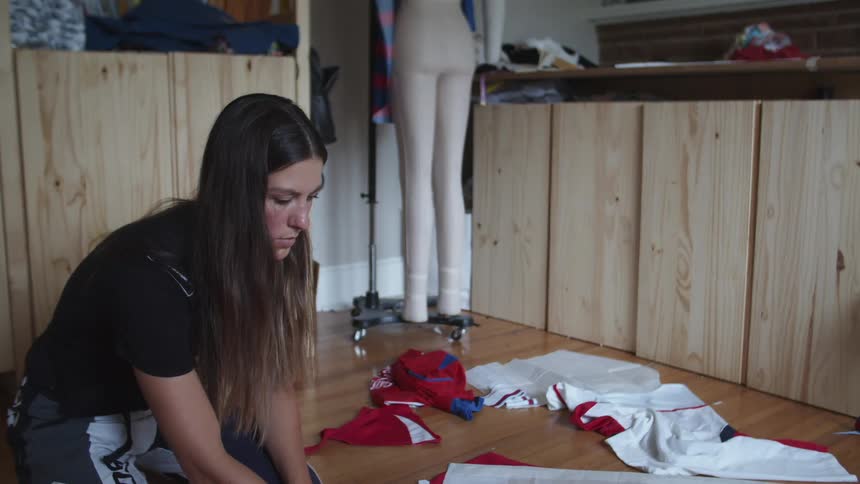 A woman sits on a hardwood floor cutting fabric with other clothes and fabric lay in piles next to her indoors