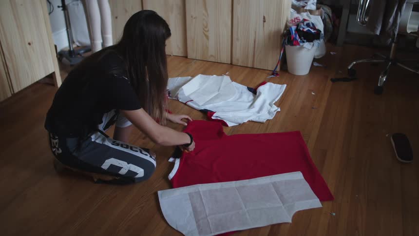 A woman with long dark hair sits on hardwood floor cutting a red shirt