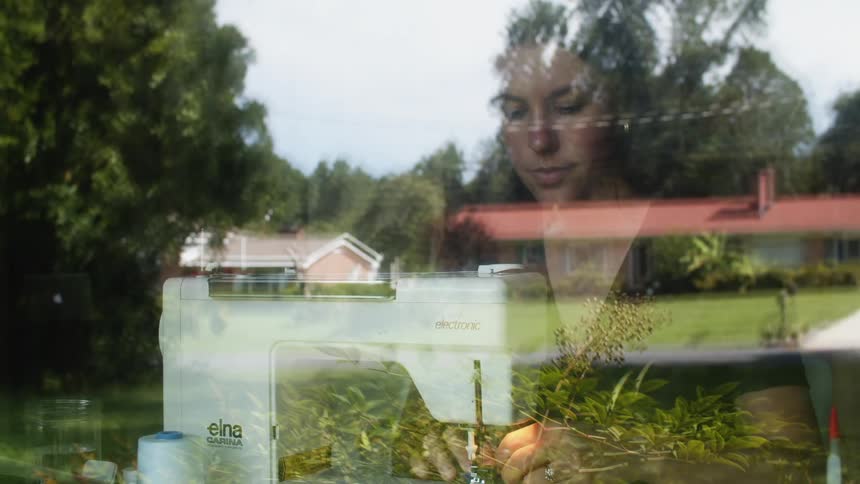 A woman is reflected in her window feeding fabric to a sewing machine