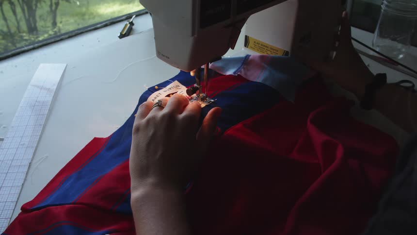 Tag being sewn onto a blue and red shirt with a sewing machine on a table indoors
