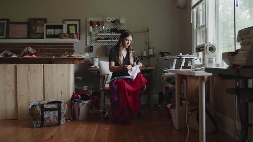 Woman sitting on a white chair in a tailoring workshop in a house adjusting the sleeves on a red and blueshirt