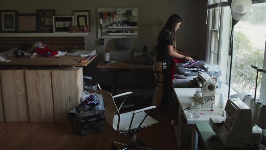 Woman sitting at a table with a sewing machine in a workshop gets up to iron the piece of clothing she is working on