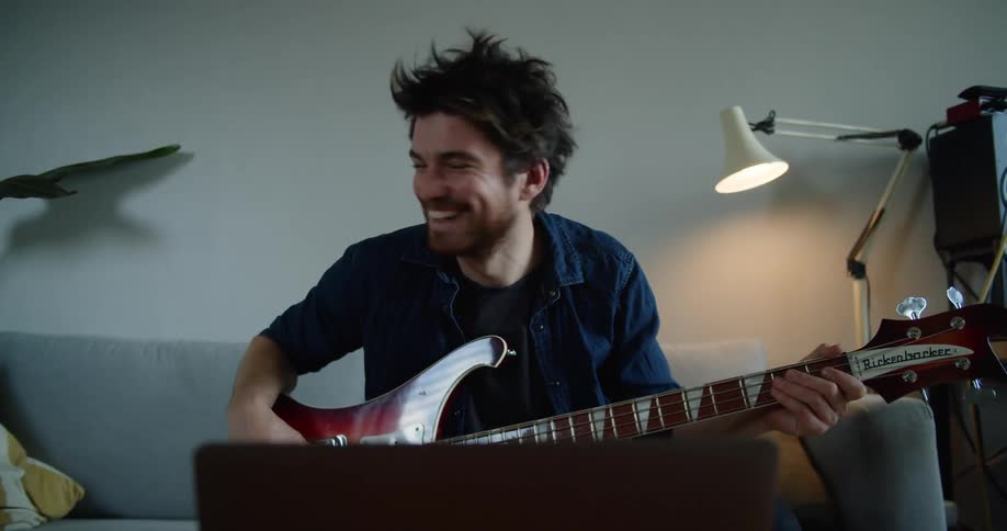 Man with a beard and a dark blue shirt plays a guitar and makes a video call on the laptop in the living room at home
