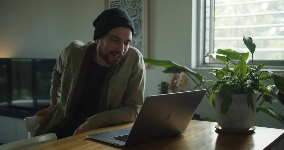 Man sitting at a desk inside of a home with a potted plant on the edge talking at his laptop screen on a video call